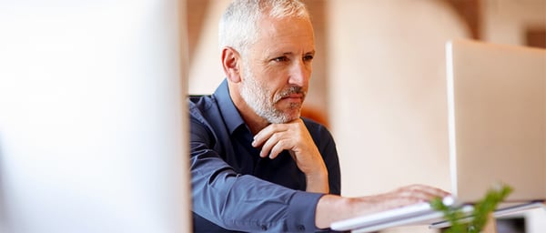 A gray-haired professional man sits at a desk, focused on a computer screen while resting his chin on his hand. Wearing a dark blue shirt, he appears thoughtful and concentrated in a warm office setting, suggesting analytical work, decision-making, or digital productivity.